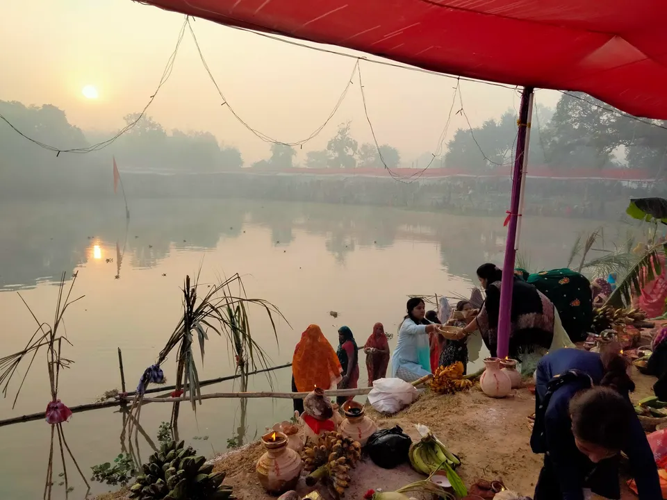 सकाळच्या धुक्यात तळ्यावरून उगवणारा सूर्य; काठावर सूप, ऊस, फळे आणि मातीची भांडी मांडून बसलेले भाविक