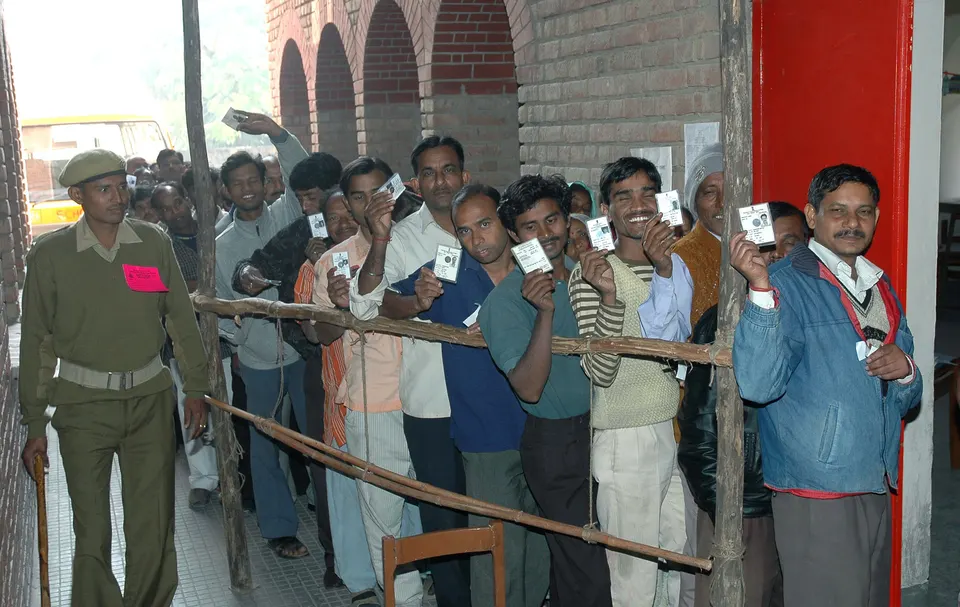 मतदान केंद्राबाहेर रांगेत उभे असलेले मतदार हातात मतदार ओळखपत्र उंचावून दाखवत आहेत, शेजारी एक पोलीस कर्मचारी उभा आहे