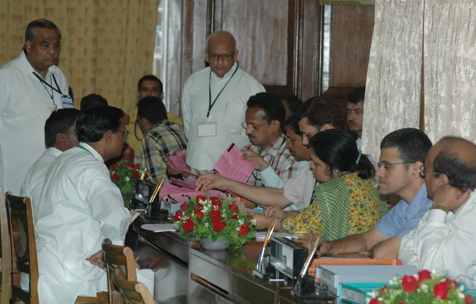 Officials seated around a counting table handle pink ballot papers while others stand behind them and watch